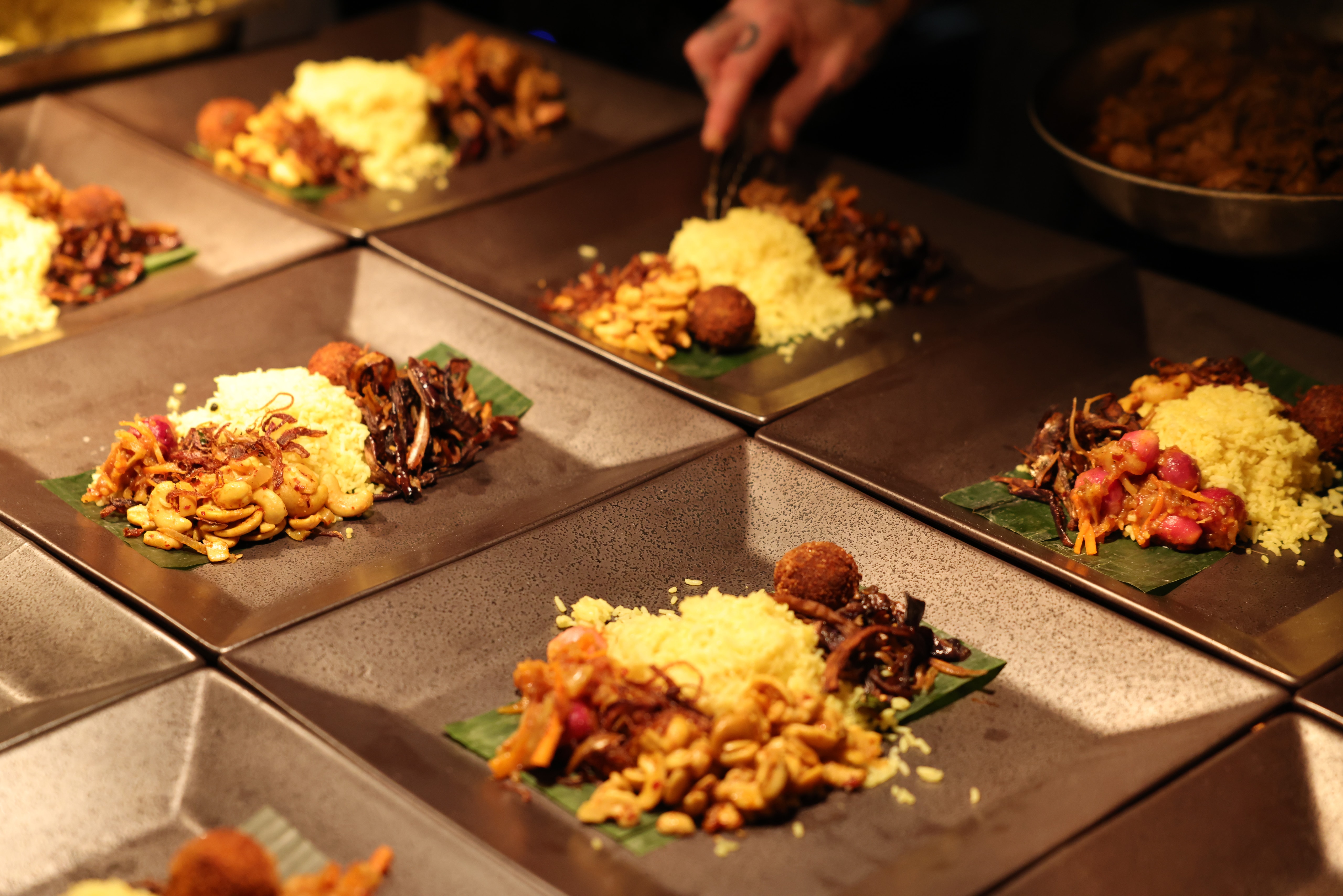 Square plates of yellow rice, fried shallots, and spiced accompaniments on banana leaves, finished by hand.